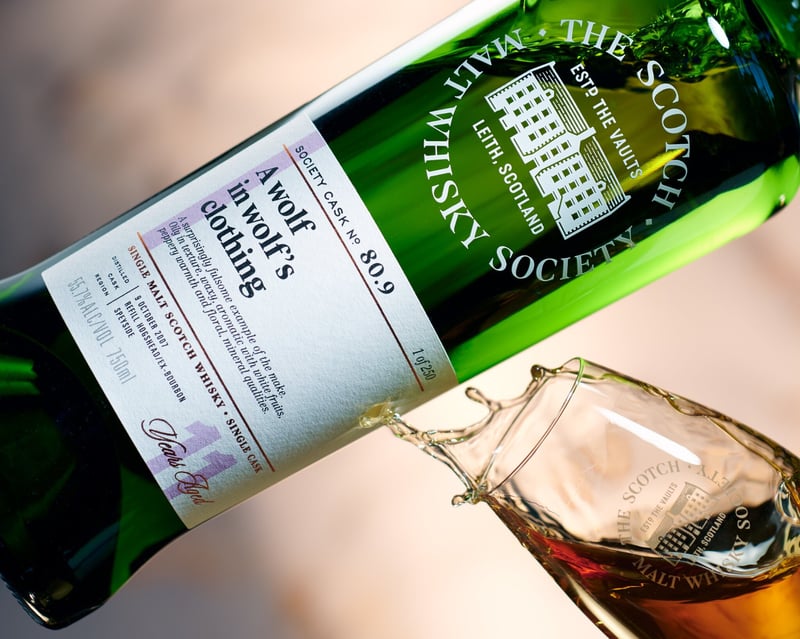 Scotch Malt Whisky Society bottle tipped mid-pour, whisky arcing into a branded glass against blurred green foliage