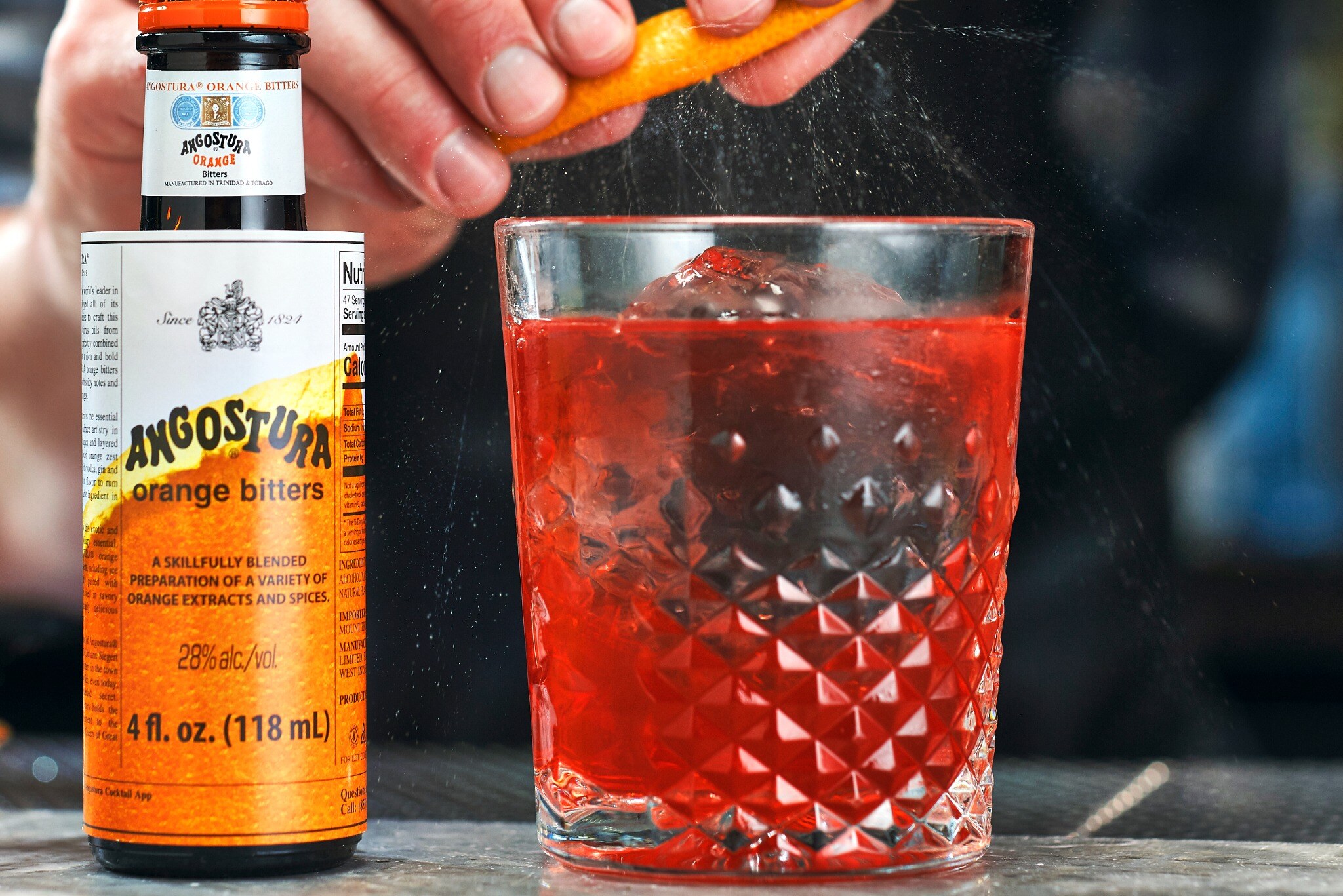A hand grates zest over a ribbed tumbler of red cocktail, a bottle of Angostura orange bitters beside it
