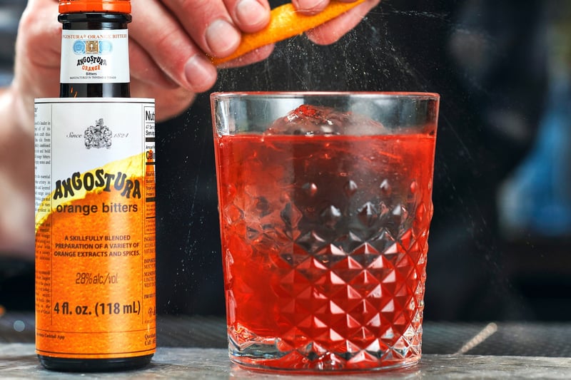 A hand grates zest over a ribbed tumbler of red cocktail, a bottle of Angostura orange bitters beside it