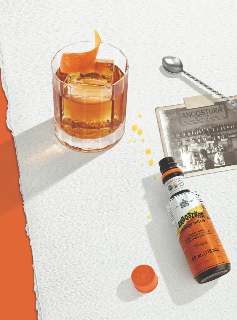 Angostura bitters bottle beside an old fashioned in a heavy tumbler with a curled orange twist, hard sunlight raking across torn white and orange paper
