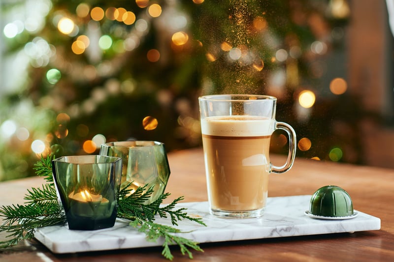 A latte in a glass mug on a marble board beside candles and greenery, festive lights blurred behind