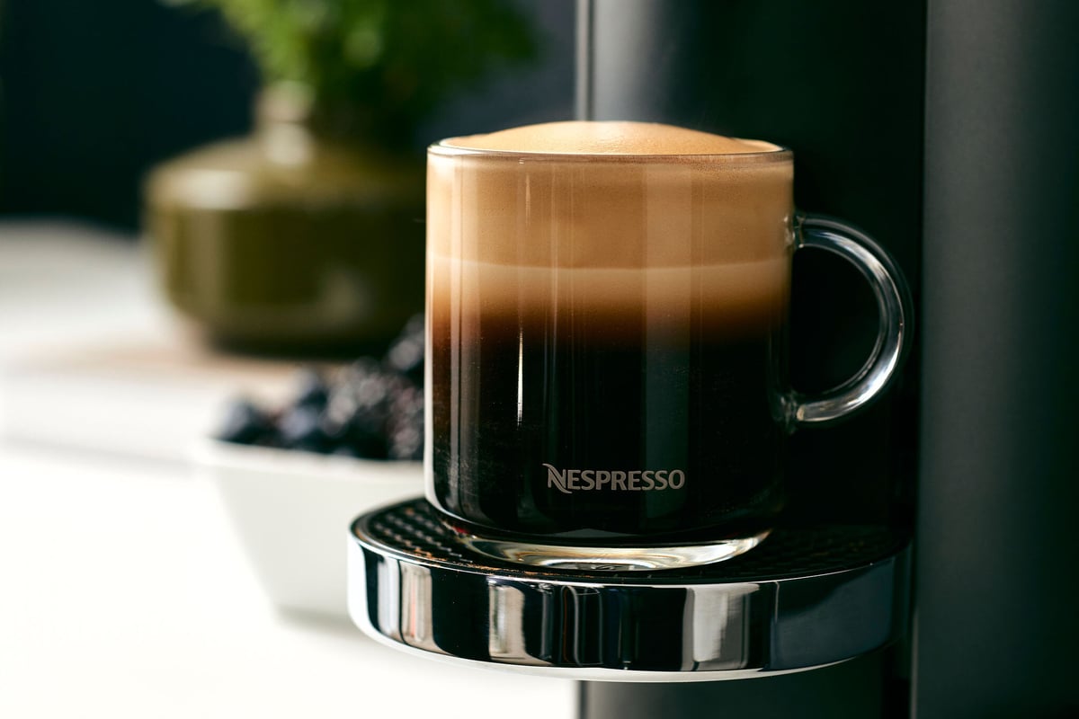 Nespresso layered latte in a clear glass mug on the machine drip tray, blackberries and a dark vase softly out of focus behind