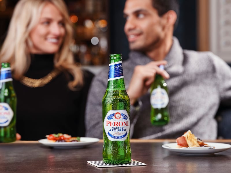 Two people talking across a bar table with Peroni Nastro Azzurro bottles and small plates between them, condensation on the green glass