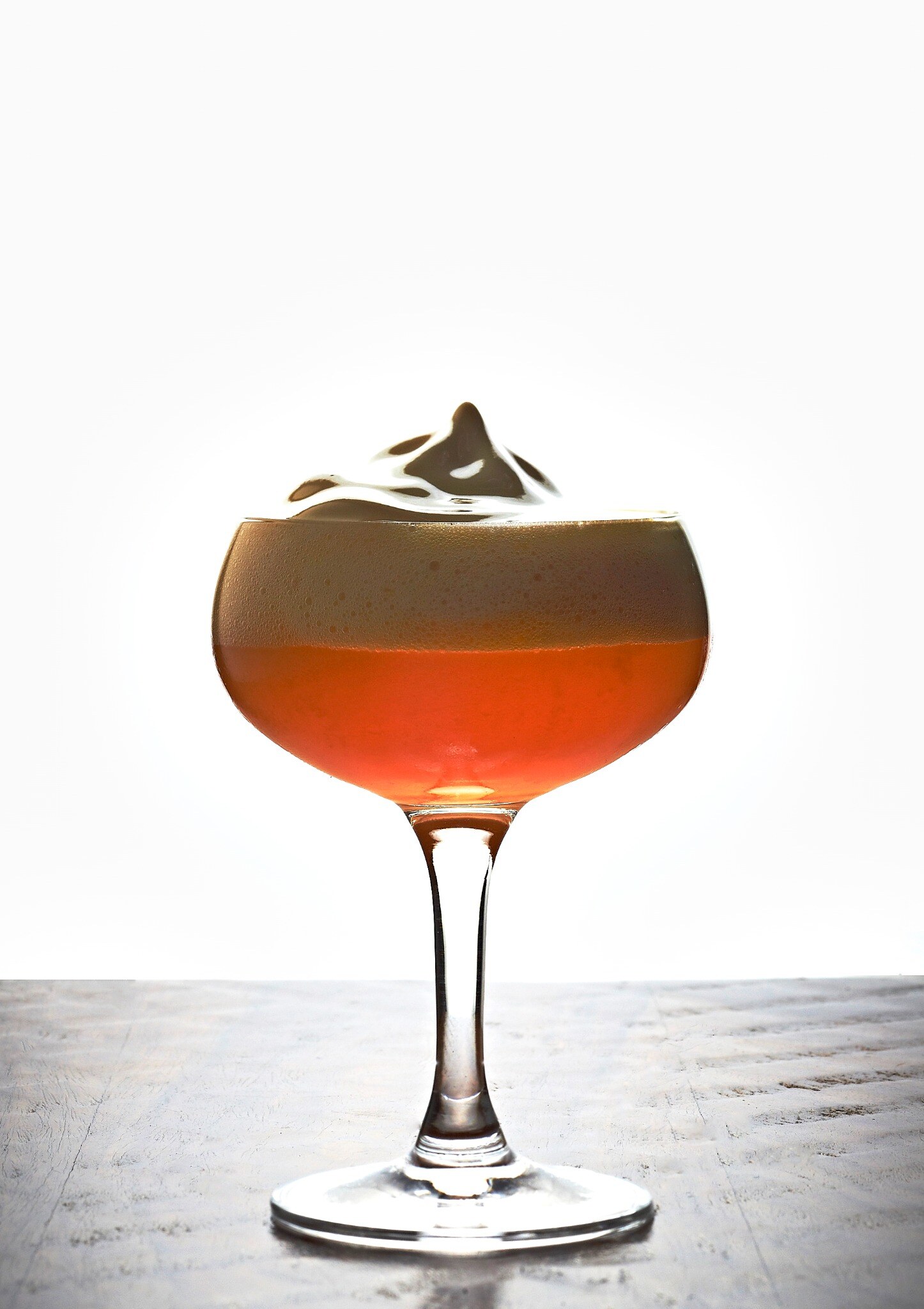Coupe glass of a dark cocktail with a thick foam cap and etched garnish, backlit on a pale sweep above a weathered wood floor