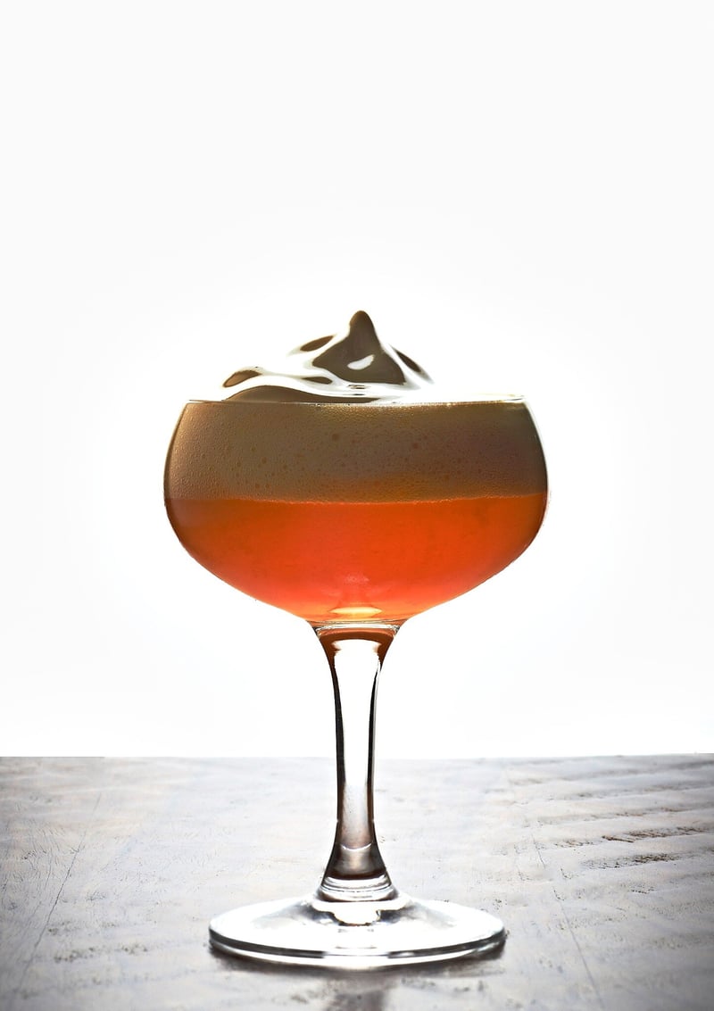 Coupe glass of a dark cocktail with a thick foam cap and etched garnish, backlit on a pale sweep above a weathered wood floor