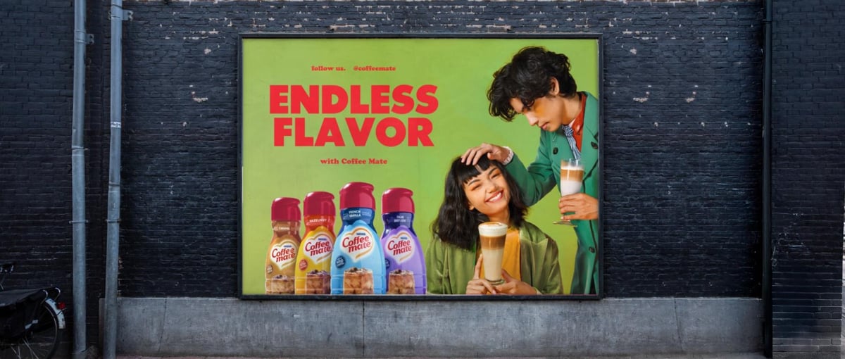 Coffee Mate billboard mounted on a dark brick wall, two people photographed mid-laugh beside a row of flavour bottles