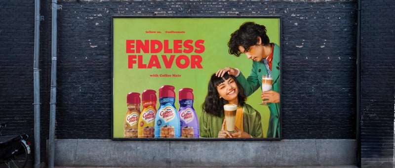 Coffee Mate billboard mounted on a dark brick wall, two people photographed mid-laugh beside a row of flavour bottles