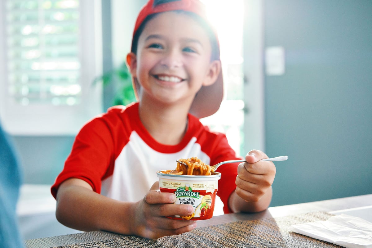 Child in a red baseball shirt and cap grinning at a kitchen counter, holding a spoon over a cup of pasta