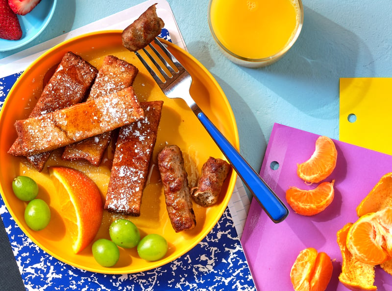 Overhead of a child's breakfast plate — French toast sticks, sausages, orange segments and grapes on bright orange melamine, juice and colored card around it