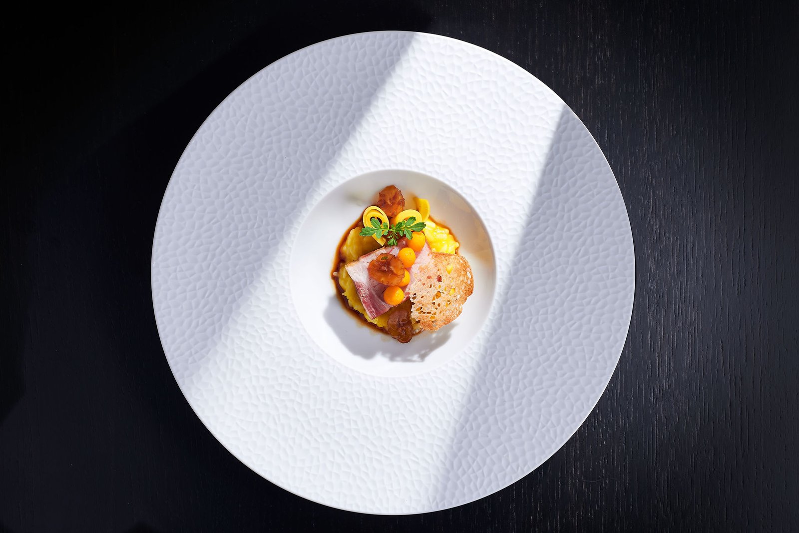 Single plated course centred on a wide white porcelain plate, hard directional light cutting across a near-black table