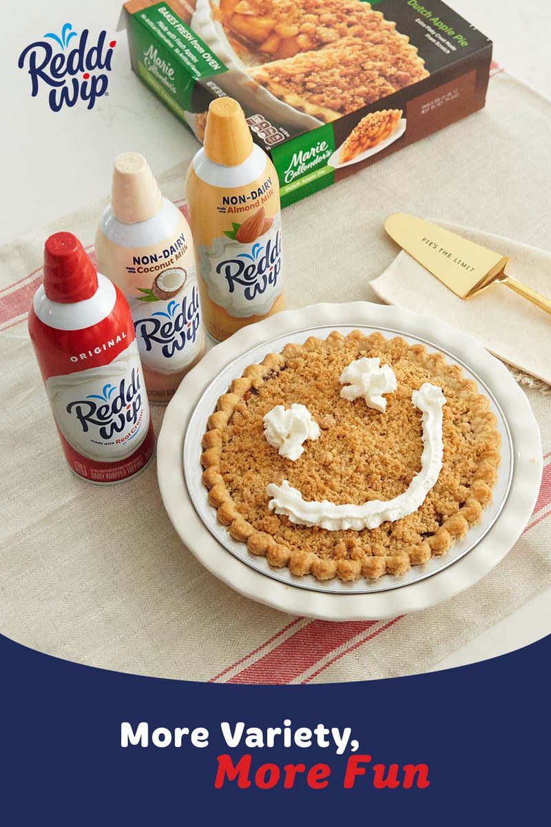 Reddi-wip cans arranged around a graham crust pie with a whipped cream smile, product carton behind on a striped cloth
