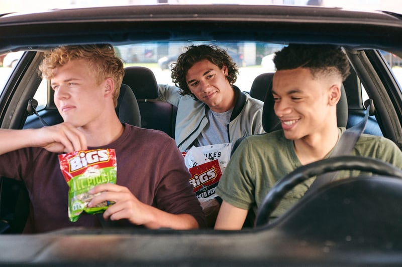 Three young men in a car share a bag of crisps, laughing through the open window