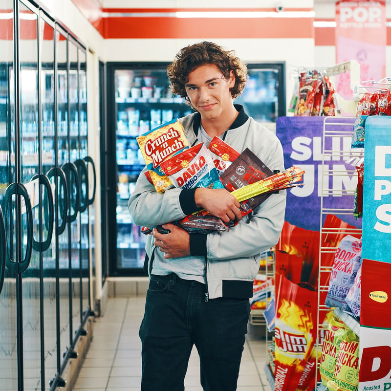 Young man in a convenience store aisle with an armload of snacks, coolers and hanging product racks receding behind him