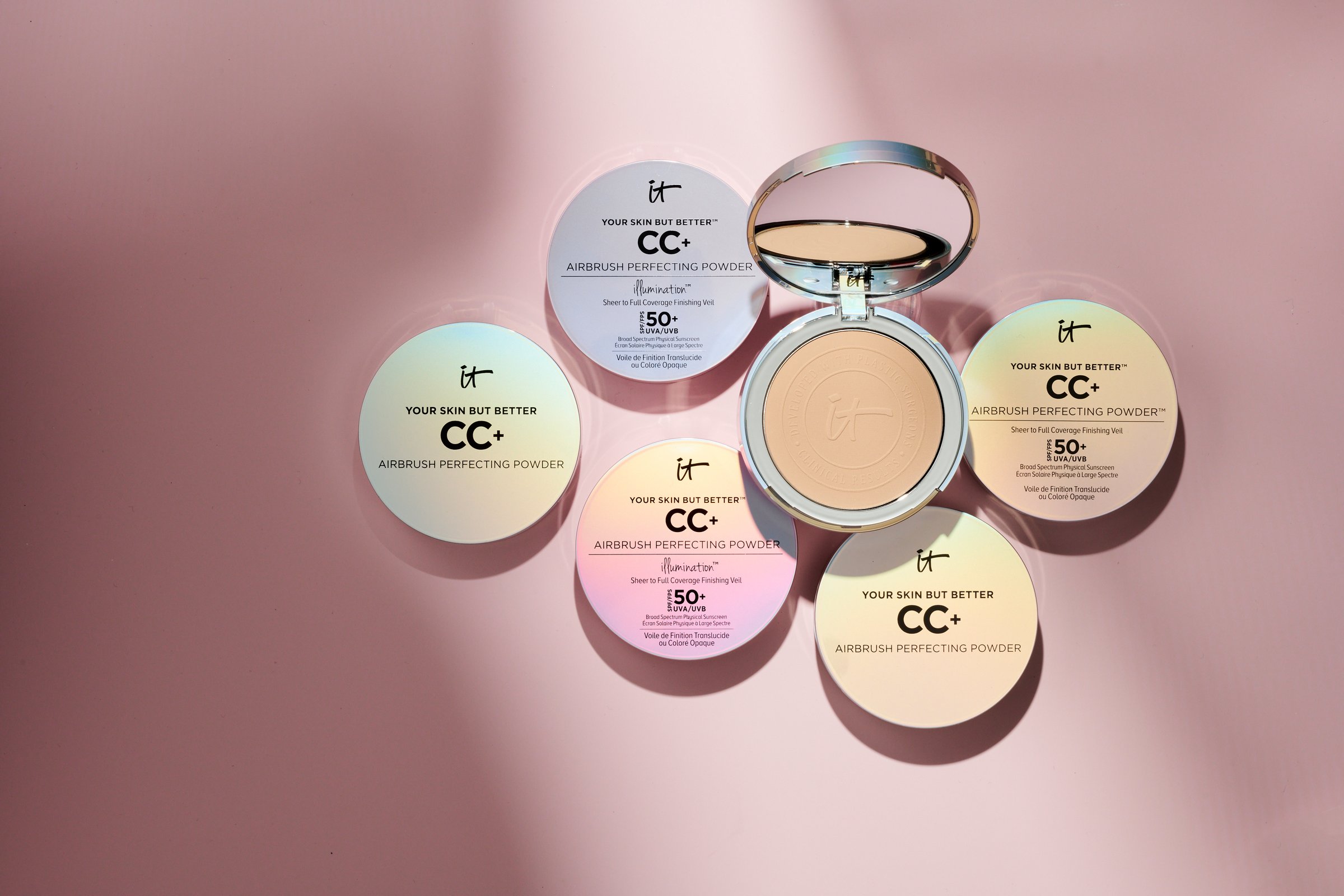 Seven IT Cosmetics CC+ compacts fanned in a circle around an open powder compact on dusty pink, long soft shadows