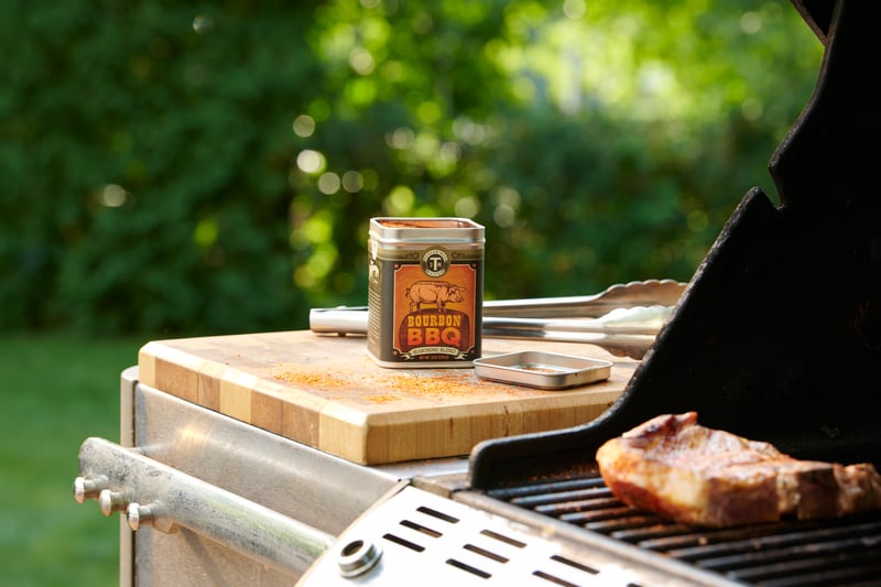 Bourbon BBQ sauce jar on a wooden board beside a grill, steak searing over the grates in dappled garden light