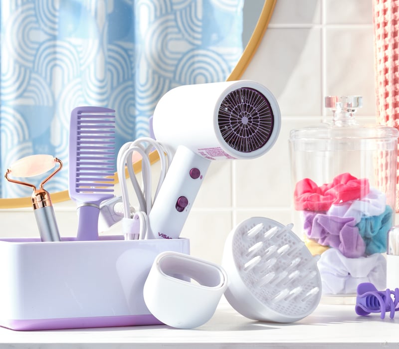 A lilac hair dryer, comb and grooming tools arranged on a pale pastel bathroom set with folded towels
