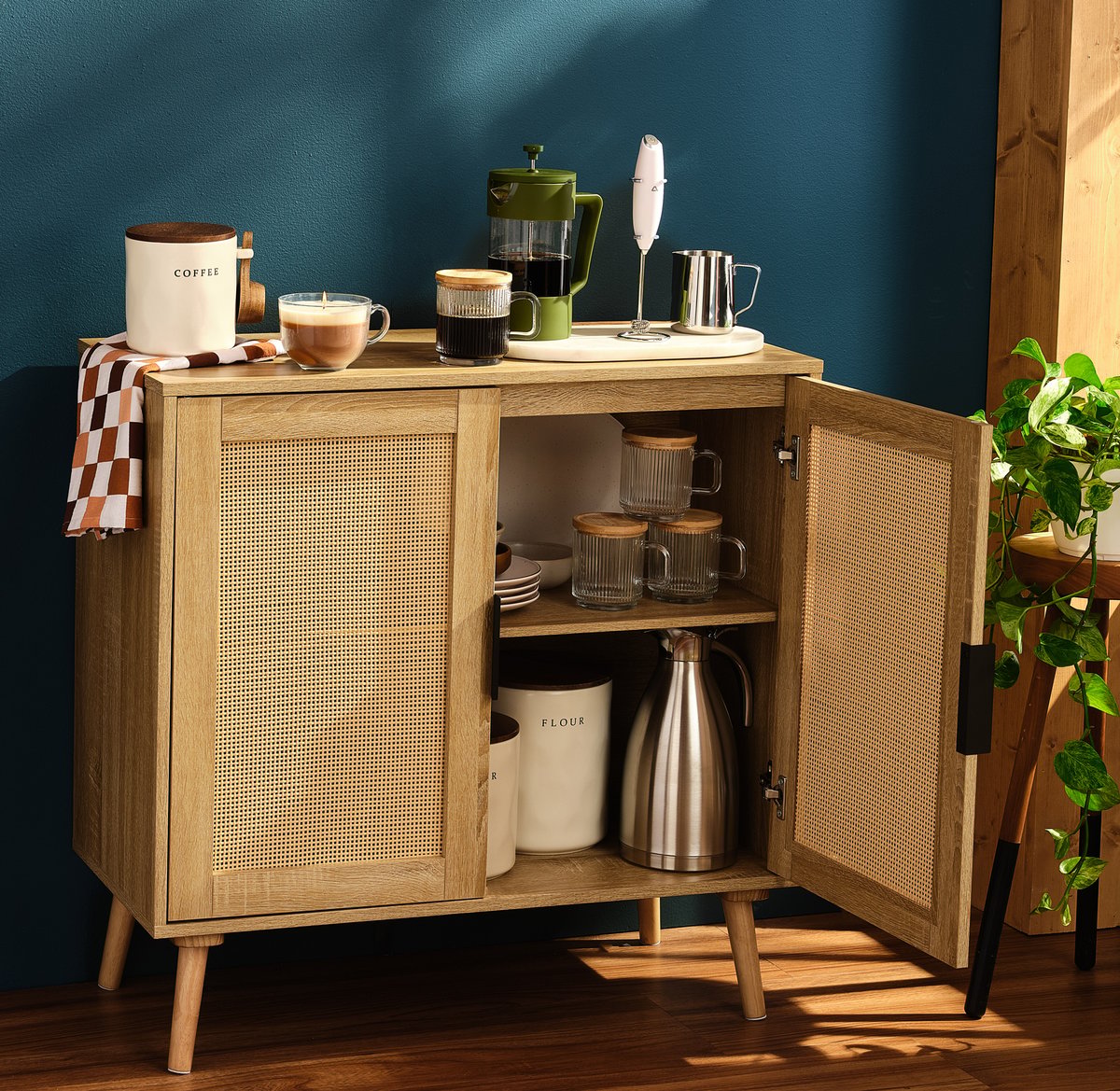 A rattan coffee cabinet standing open with an espresso machine, mugs and a kettle inside, against a teal wall