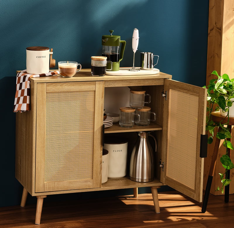 A rattan coffee cabinet standing open with an espresso machine, mugs and a kettle inside, against a teal wall