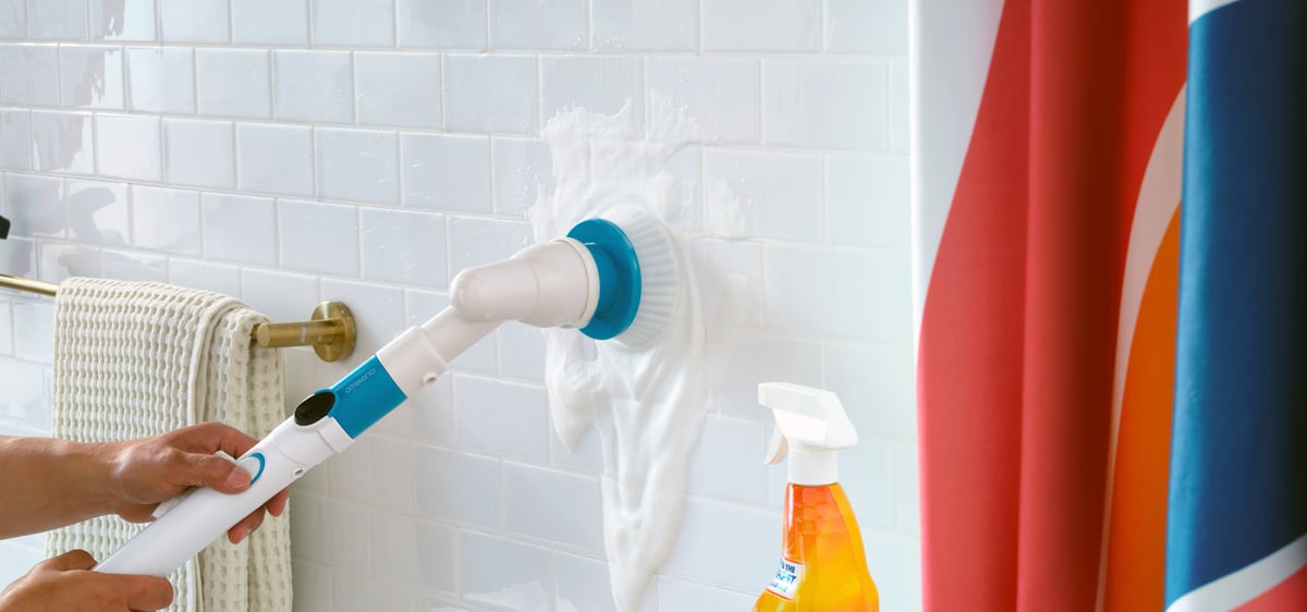 A gloved hand scrubs white tile with an electric brush beside cleaning sprays