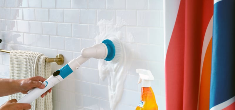 A gloved hand scrubs white tile with an electric brush beside cleaning sprays