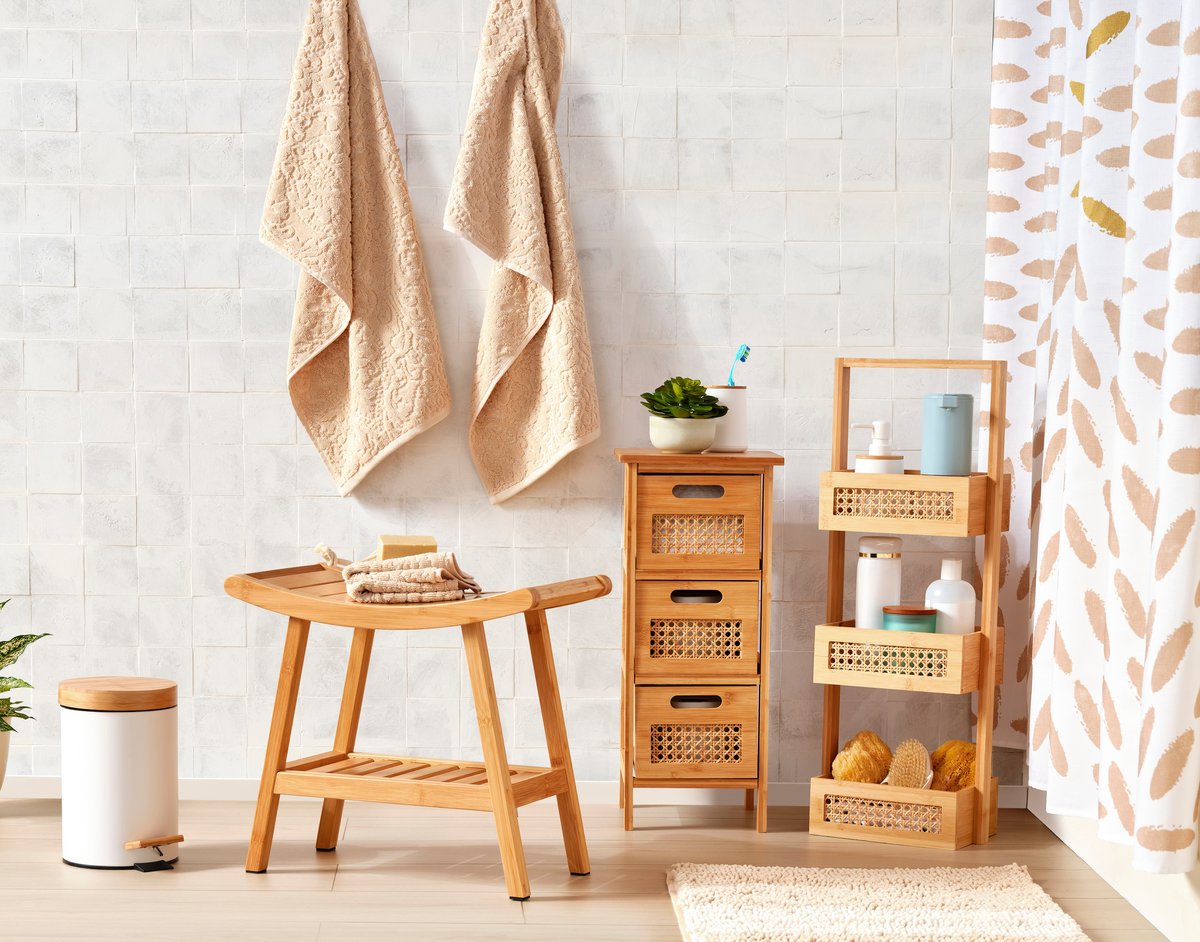 A bathroom corner with a wooden stool, tiered rattan shelving and hanging towels against white brick