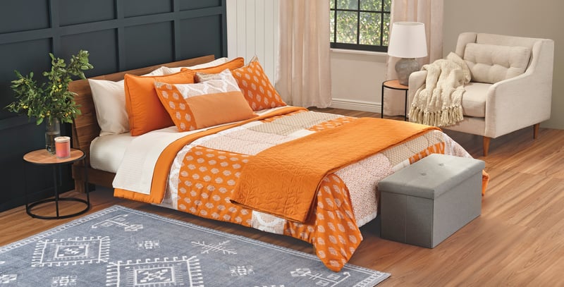 A bedroom styled in orange bedding with a grey ottoman and armchair on a patterned rug