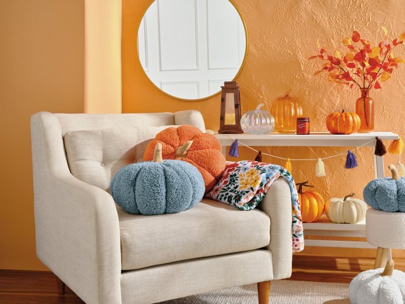 A cream armchair styled with knitted pumpkin cushions against a warm orange wall, autumn foliage on a side table