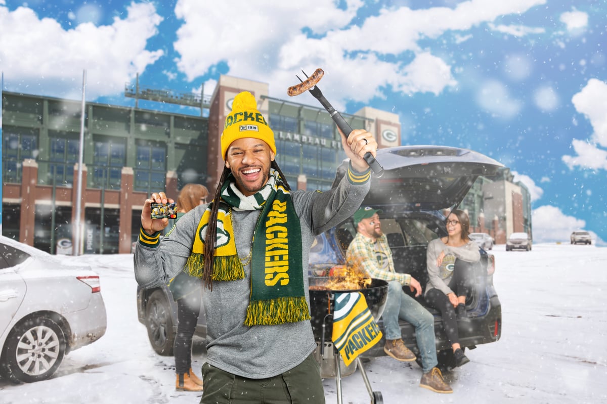 Packers supporters celebrating in falling snow outside the stadium, one raising a can, scarves and knit hats in team colors