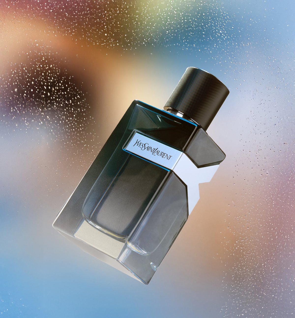 A dark glass Yves Saint Laurent fragrance bottle tilted against an iridescent blue and gold backdrop scattered with droplets