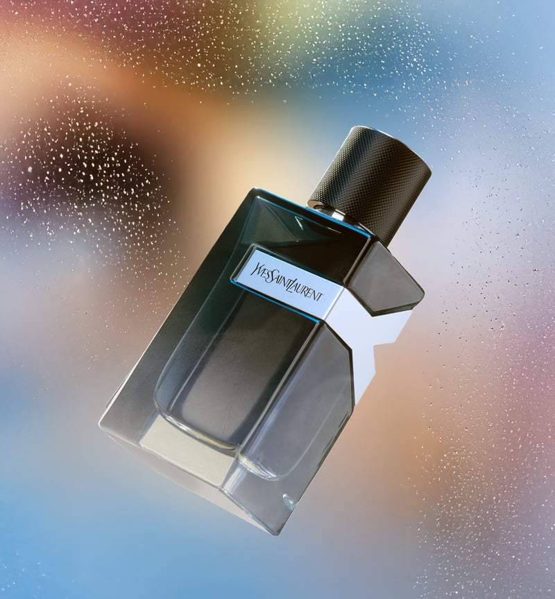 A dark glass Yves Saint Laurent fragrance bottle tilted against an iridescent blue and gold backdrop scattered with droplets