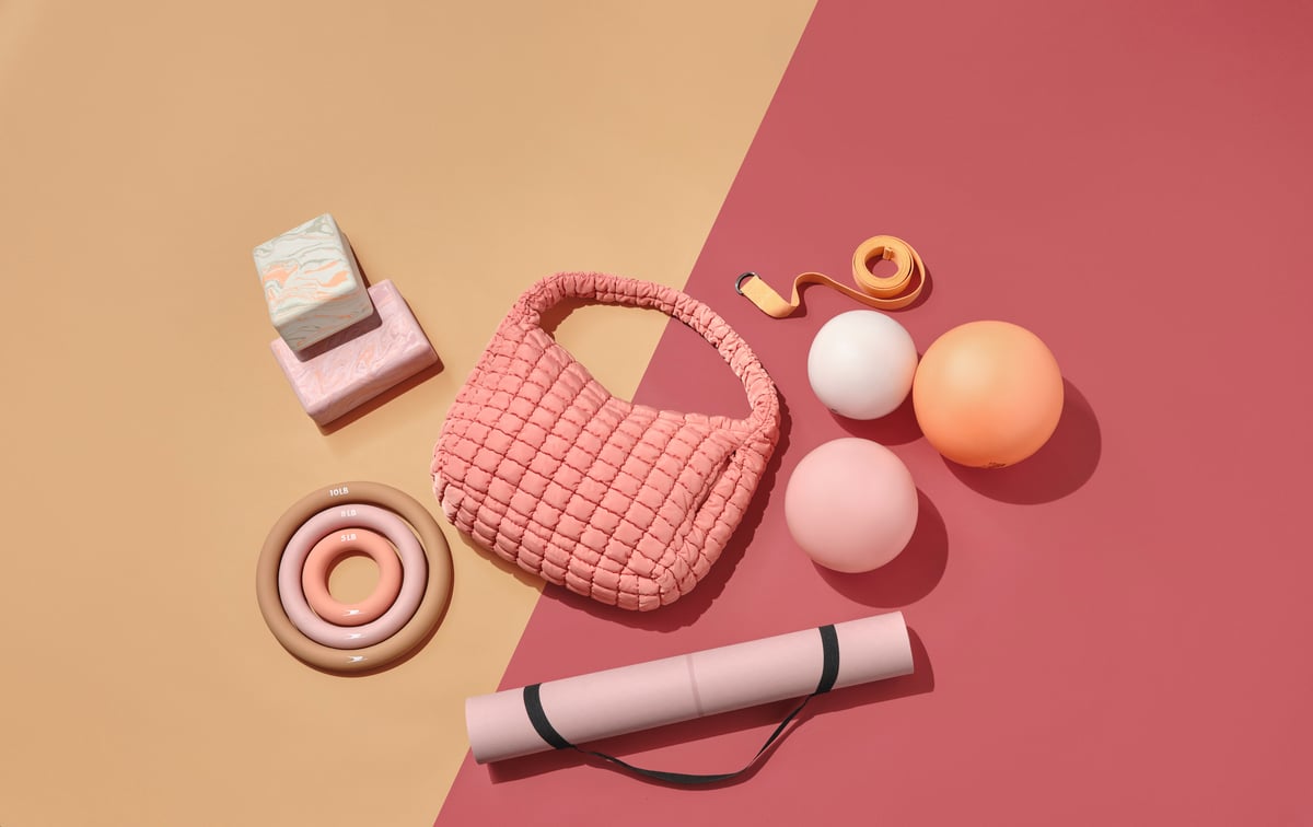 Overhead flat lay on split peach and pink card — quilted bag, yoga mat, candle and pastel spheres arranged with even spacing