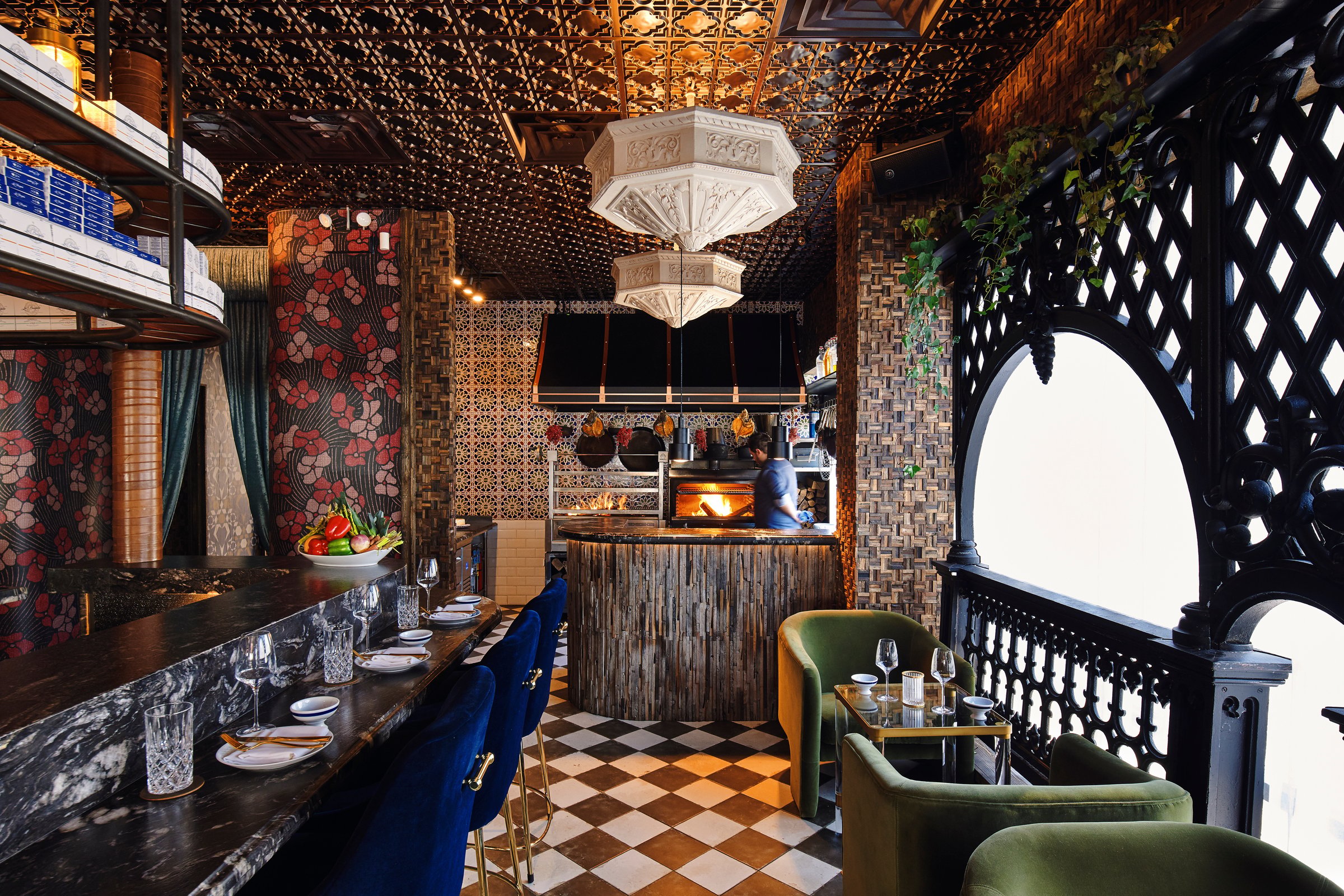 An ornate restaurant interior with a beaded chandelier, checkerboard floor and green velvet banquettes under a tiled arch