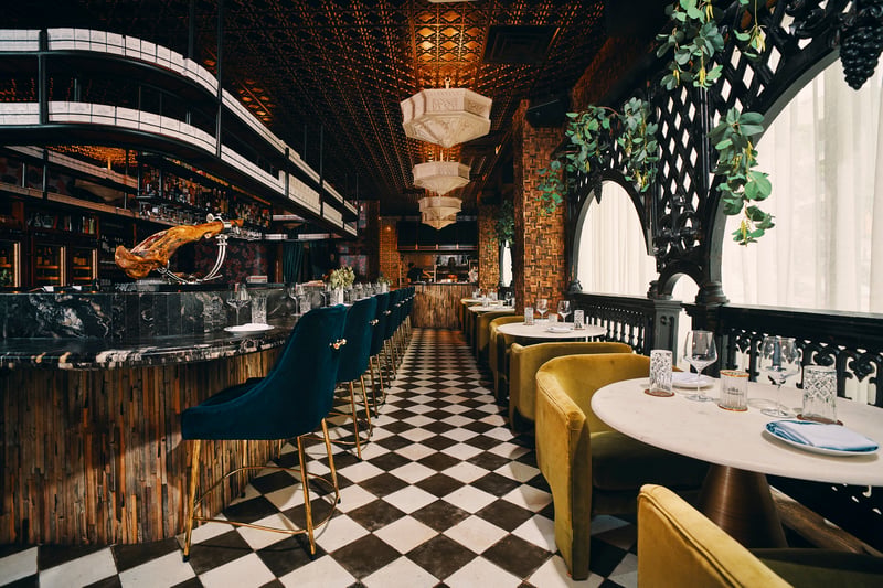 Restaurant dining room with checkerboard floor, teal and mustard banquettes, brass chandeliers and a mirrored bar behind
