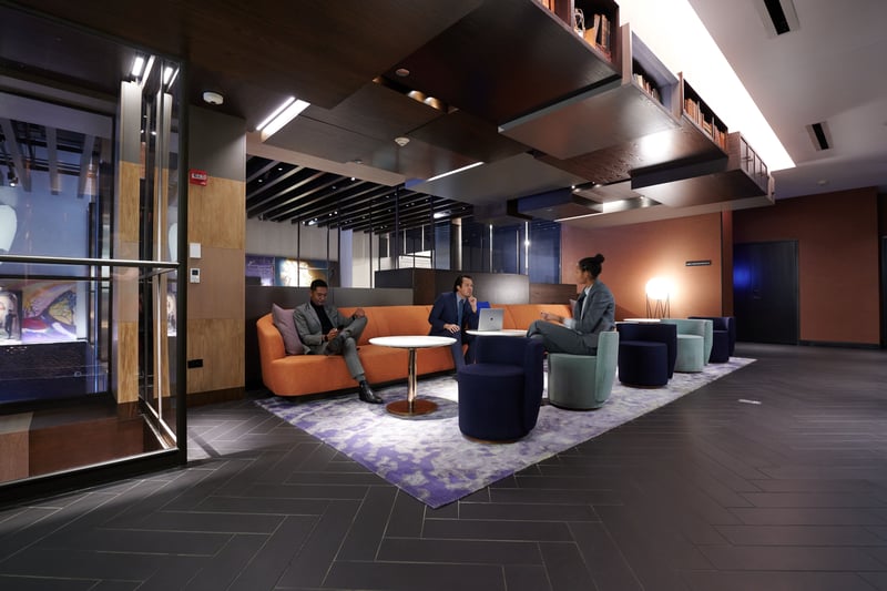 A modern office lounge with orange banquette seating and dark ceilings under tall windows