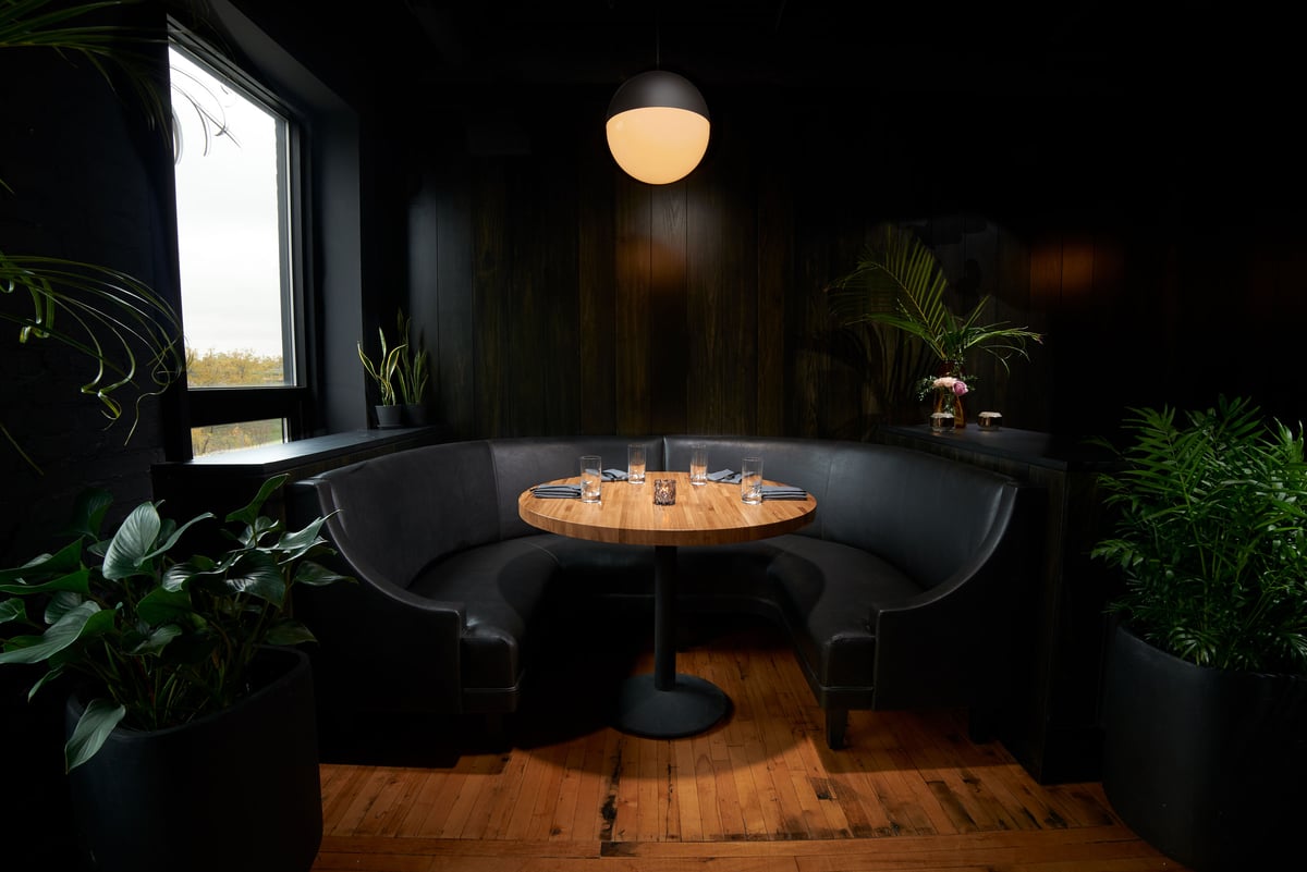 A dark dining room with a curved booth under a single pendant light, plants either side