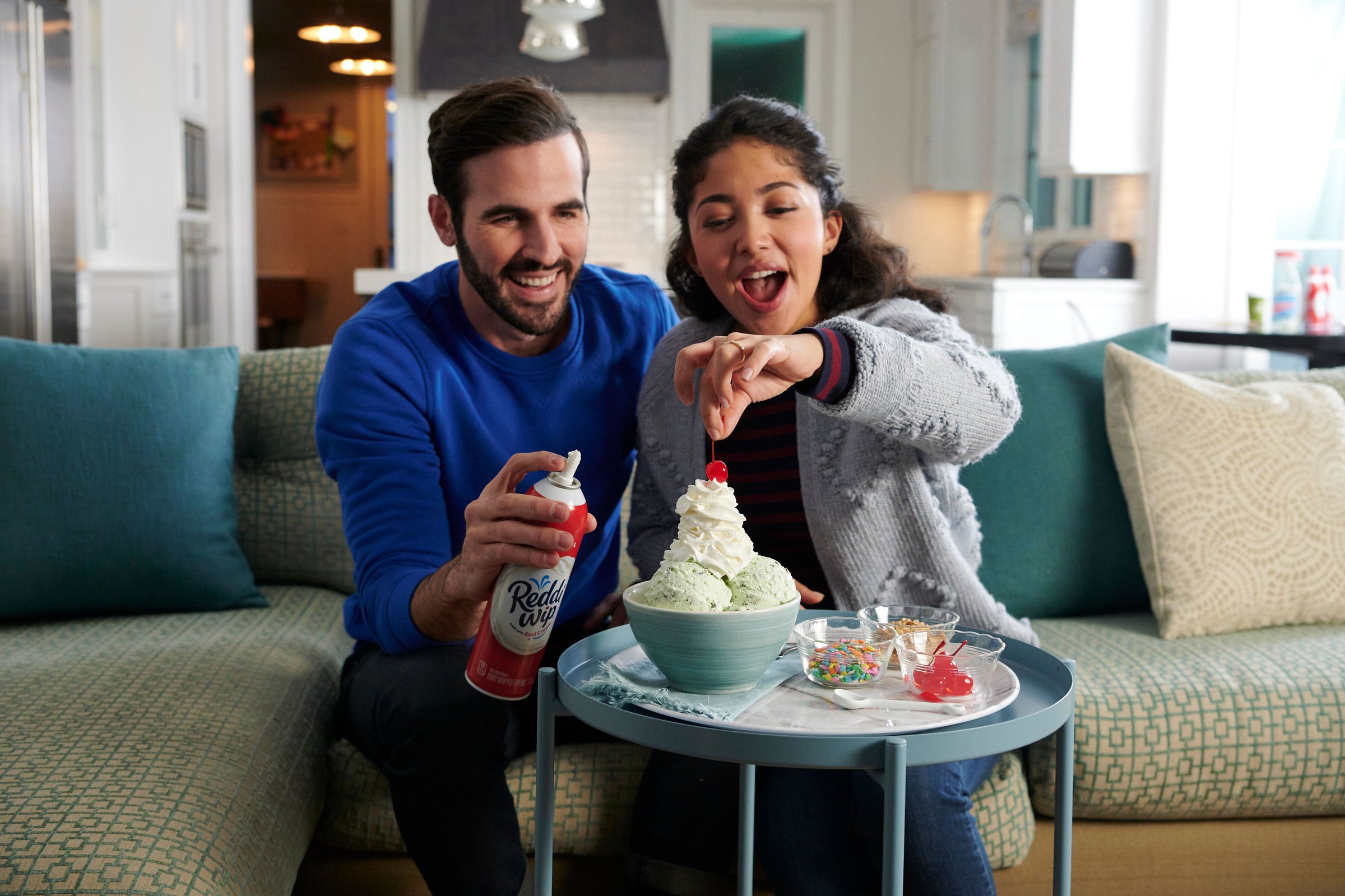 A couple on a sofa laugh as they spray whipped cream onto a dessert