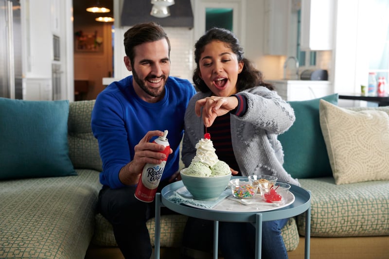 A couple on a sofa laugh as they spray whipped cream onto a dessert