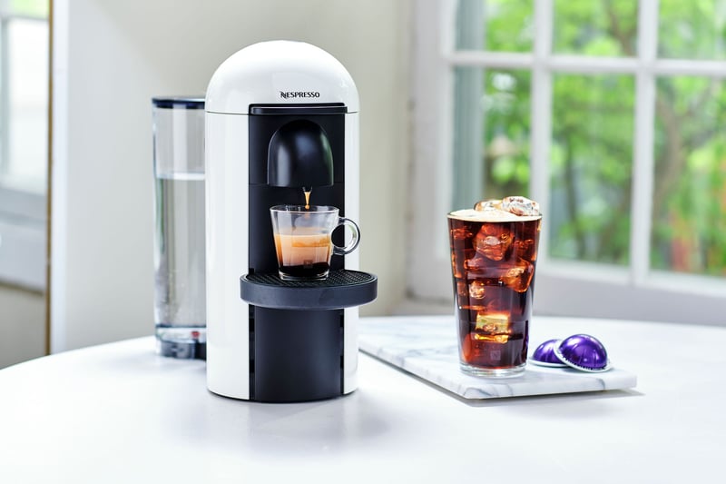 A Nespresso machine on a white counter with a filled cup, capsules and an iced coffee beside it