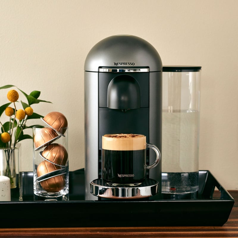 A Nespresso Vertuo machine with a filled cup on a dark tray, capsules and a citrus plant beside it