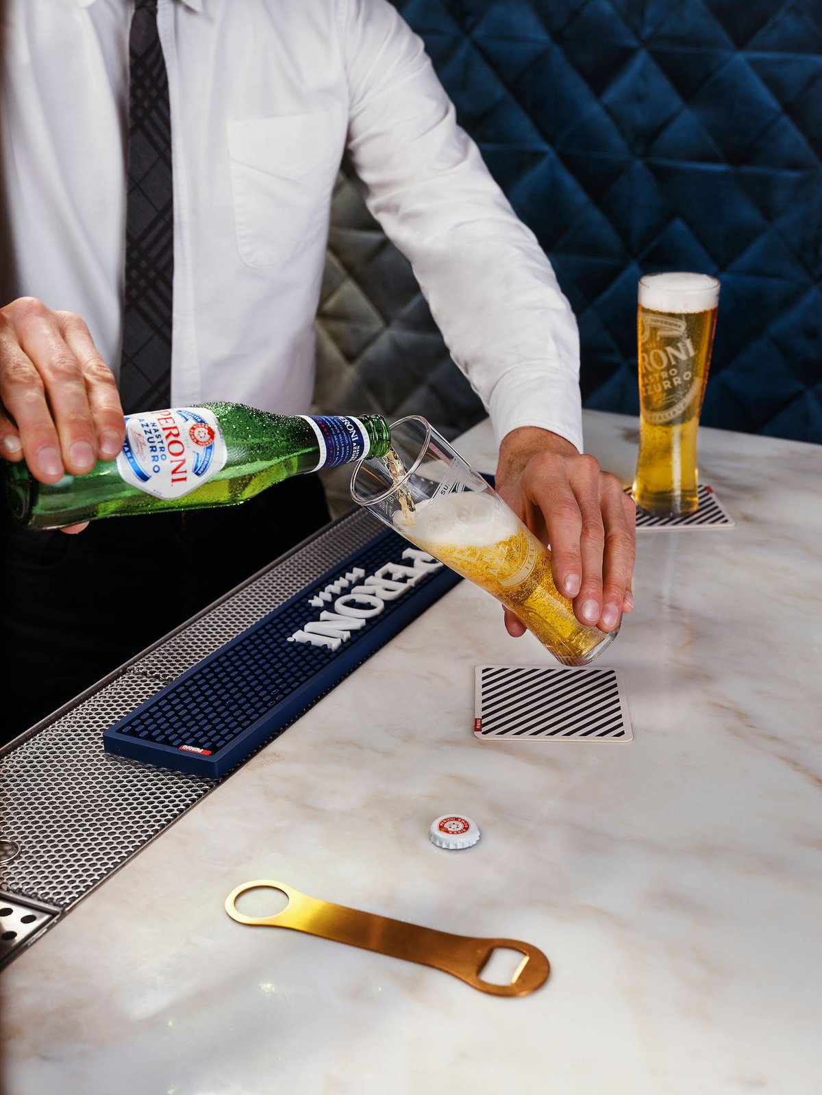 A bartender pours a bottle of Peroni into a tall glass on a marble counter