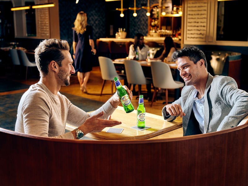 Two men talk over bottles of lager at a curved wooden bar