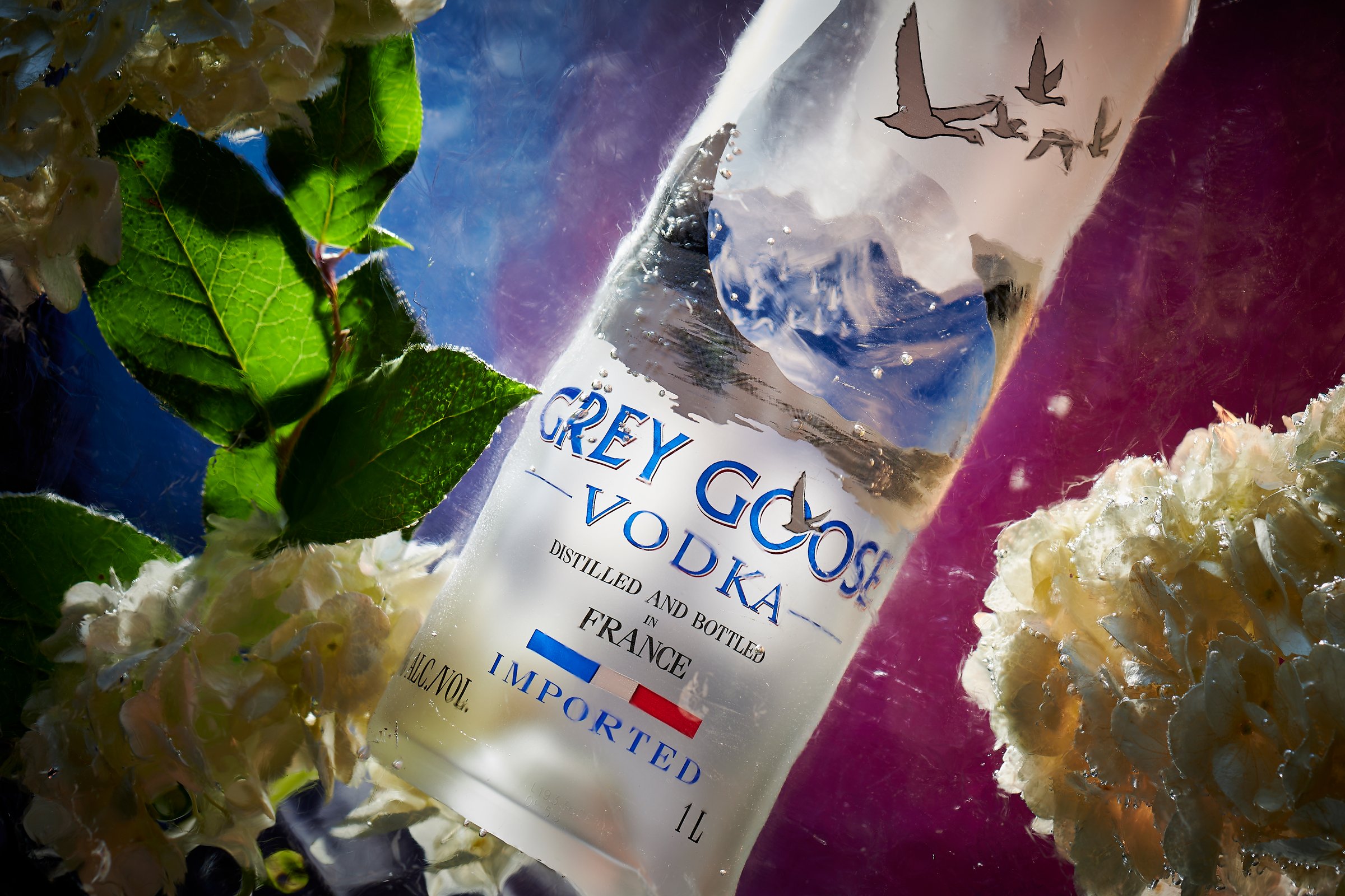 A Grey Goose vodka bottle lying among mint leaves and pale flowers under cool light