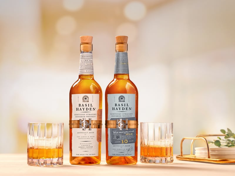 Two Basil Hayden bottles on a pale counter with cut-glass tumblers of bourbon either side