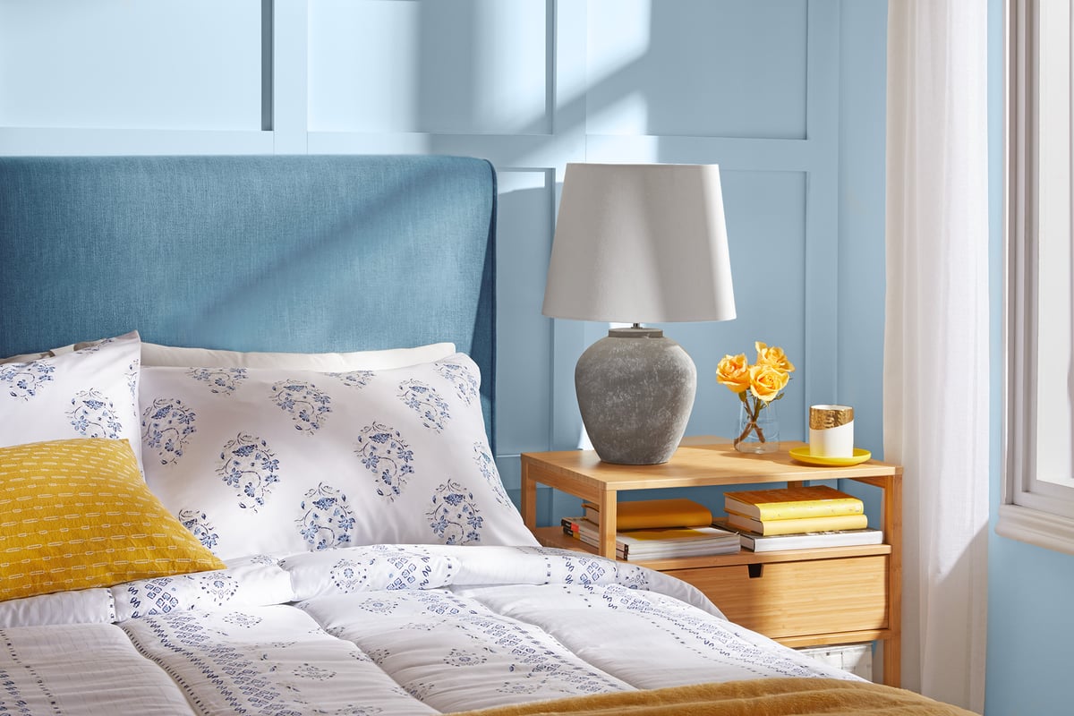 A bed made up in patterned blue and white linen with a mustard cushion beside a bright window