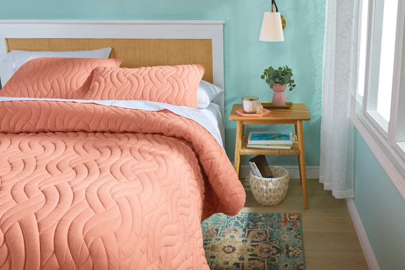 A bedroom with coral quilted bedding and a small wooden side table against a mint wall