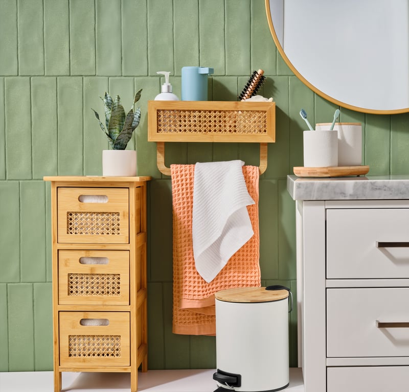 A bathroom corner with rattan drawers, a round mirror and folded towels against a green panelled wall
