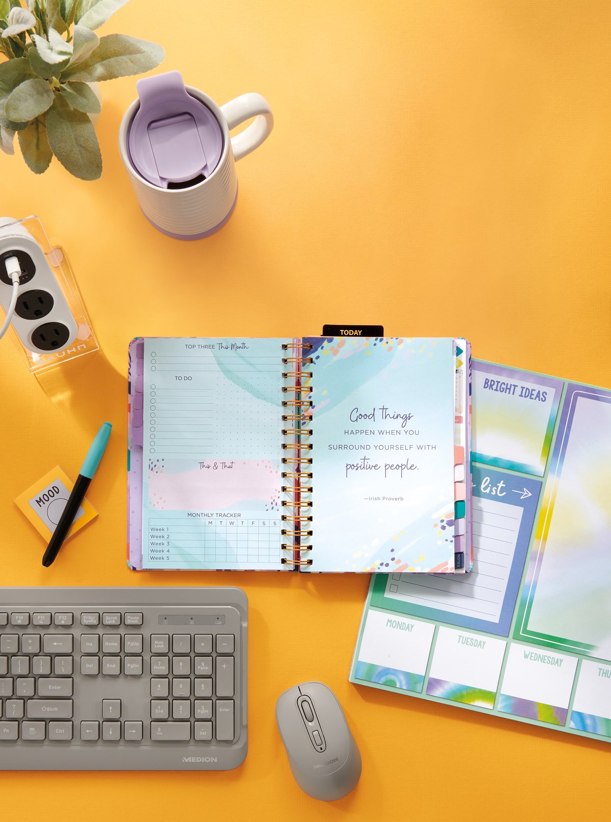 An overhead desk scene with an open planner, keyboard, mug and stationery on a yellow ground
