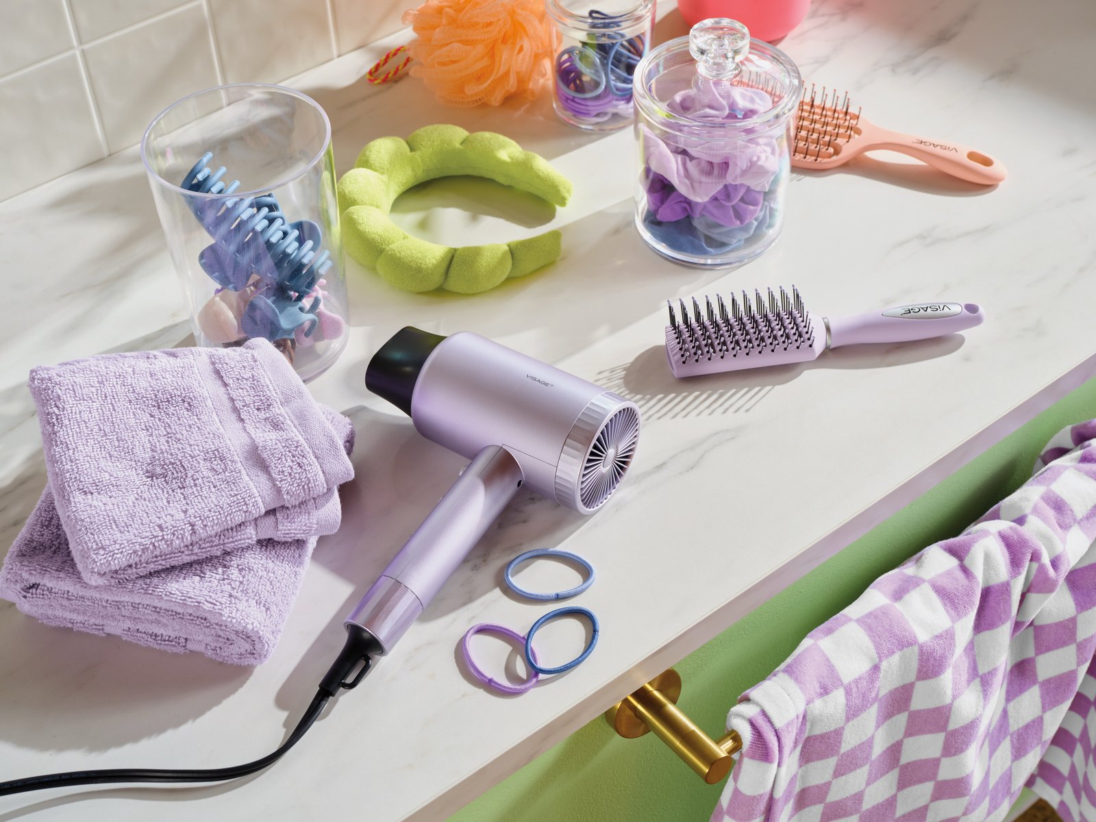 Lilac hair dryer, round brushes, scrunchies and folded towels arranged on a white marble bathroom counter in soft daylight