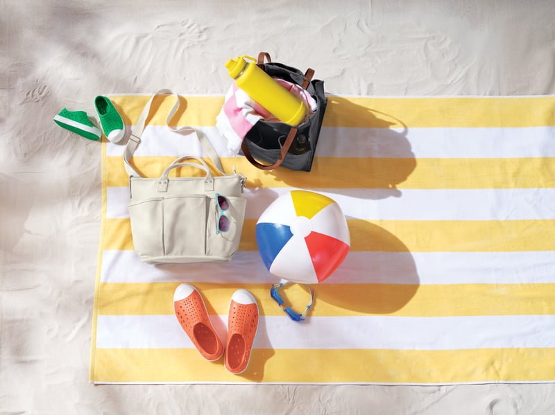An overhead beach scene with a striped towel, sandals, a beach ball and a cool bag
