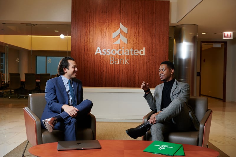 Two men in suits seated in low armchairs mid-conversation beneath the Associated Bank wordmark on a walnut wall, laptop and folder on the table between them